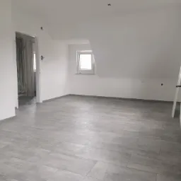 Pusty pokój na poddaszu z szarymi panelami podłogowymi imitującymi płytki, świeżo pomalowane białe ściany, okno z białymi ramami i widoczne gniazdko elektryczne.