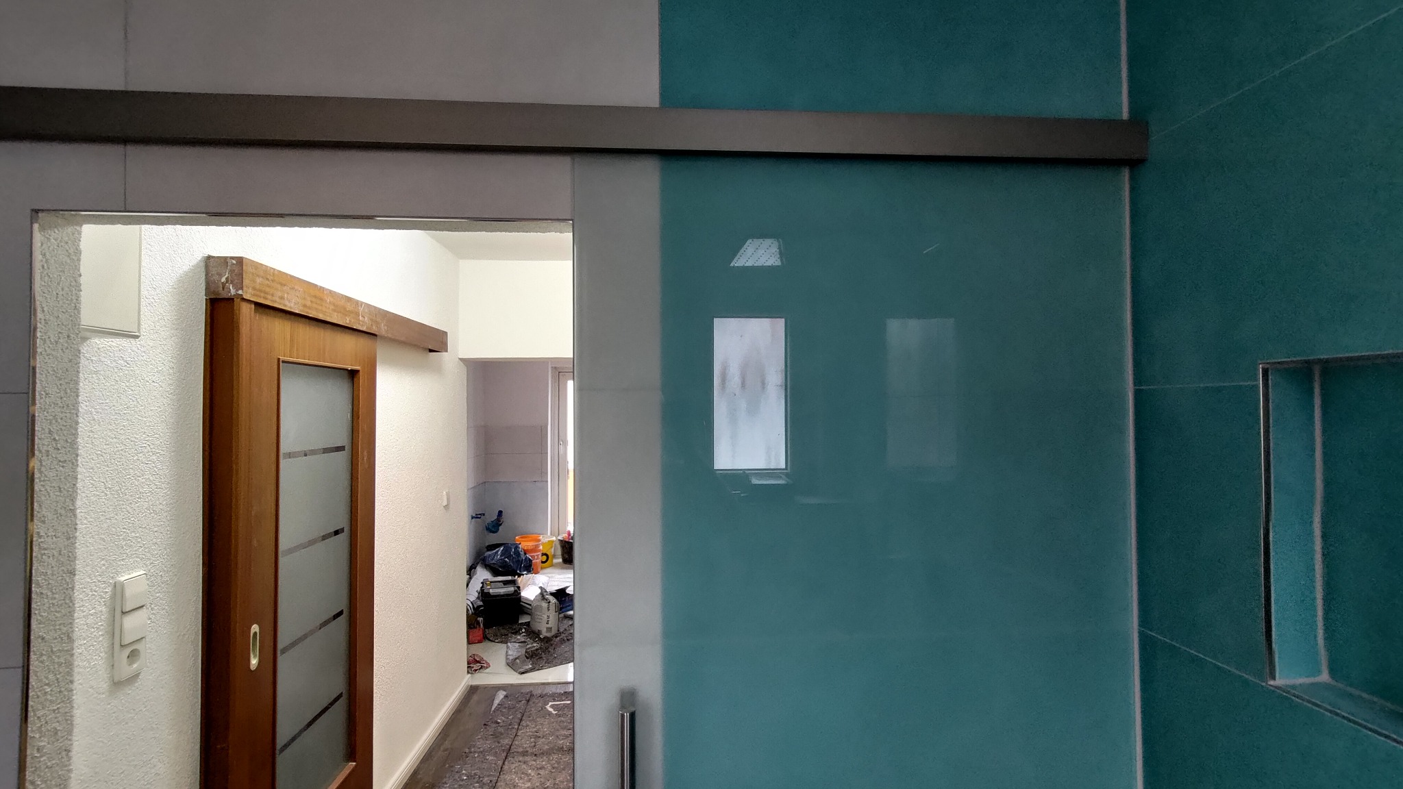 Zamontowane przesuwne drzwi łazienkowe z drewnianą ramą i matowym szkłem, widoczne turkusowe płytki na ścianie oraz fragment wnętrza łazienki z widocznymi narzędziami i materiałami budowlanymi.