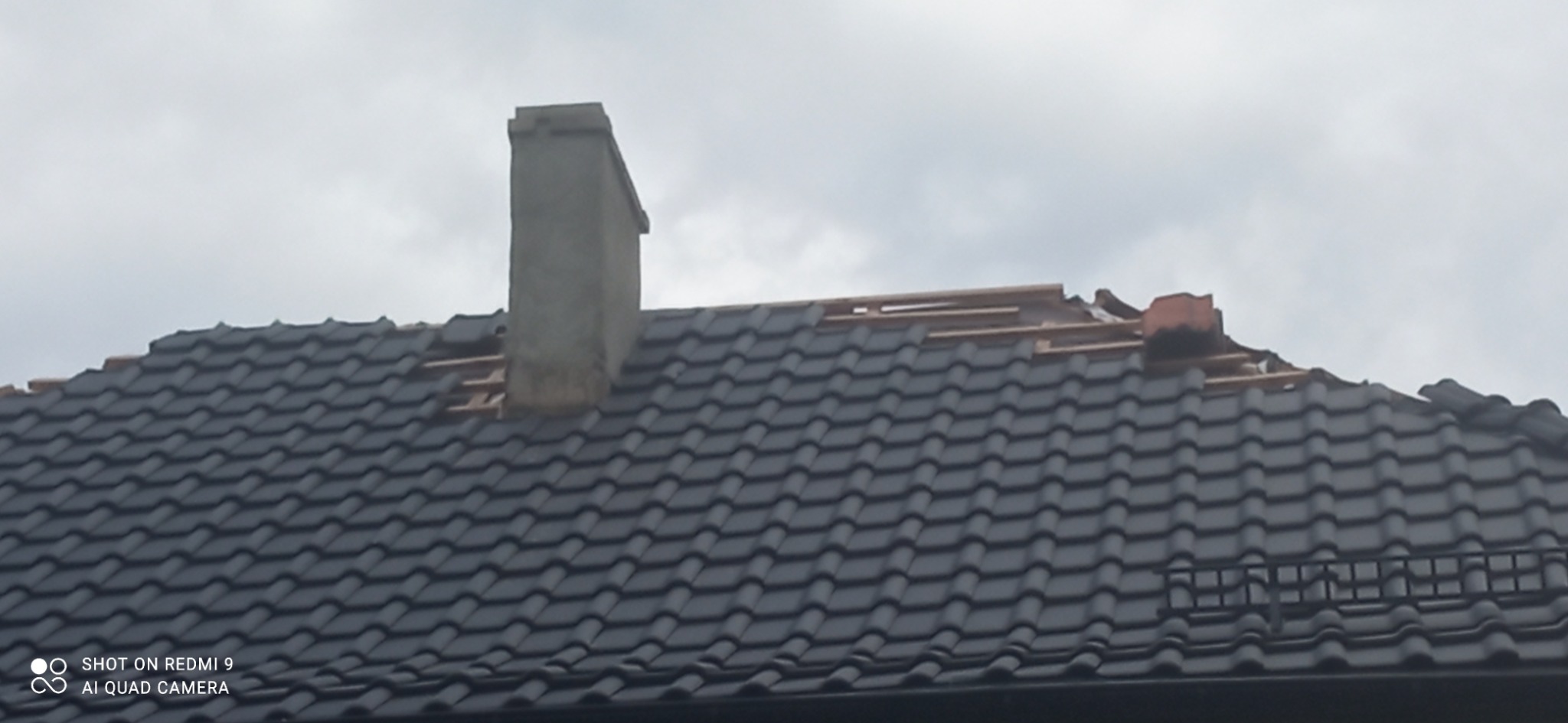 Fragment dachu z czarną dachówką, częściowo zdjętą, z widocznym kominem i elementami konstrukcji drewnianej pod spodem, na tle pochmurnego nieba.