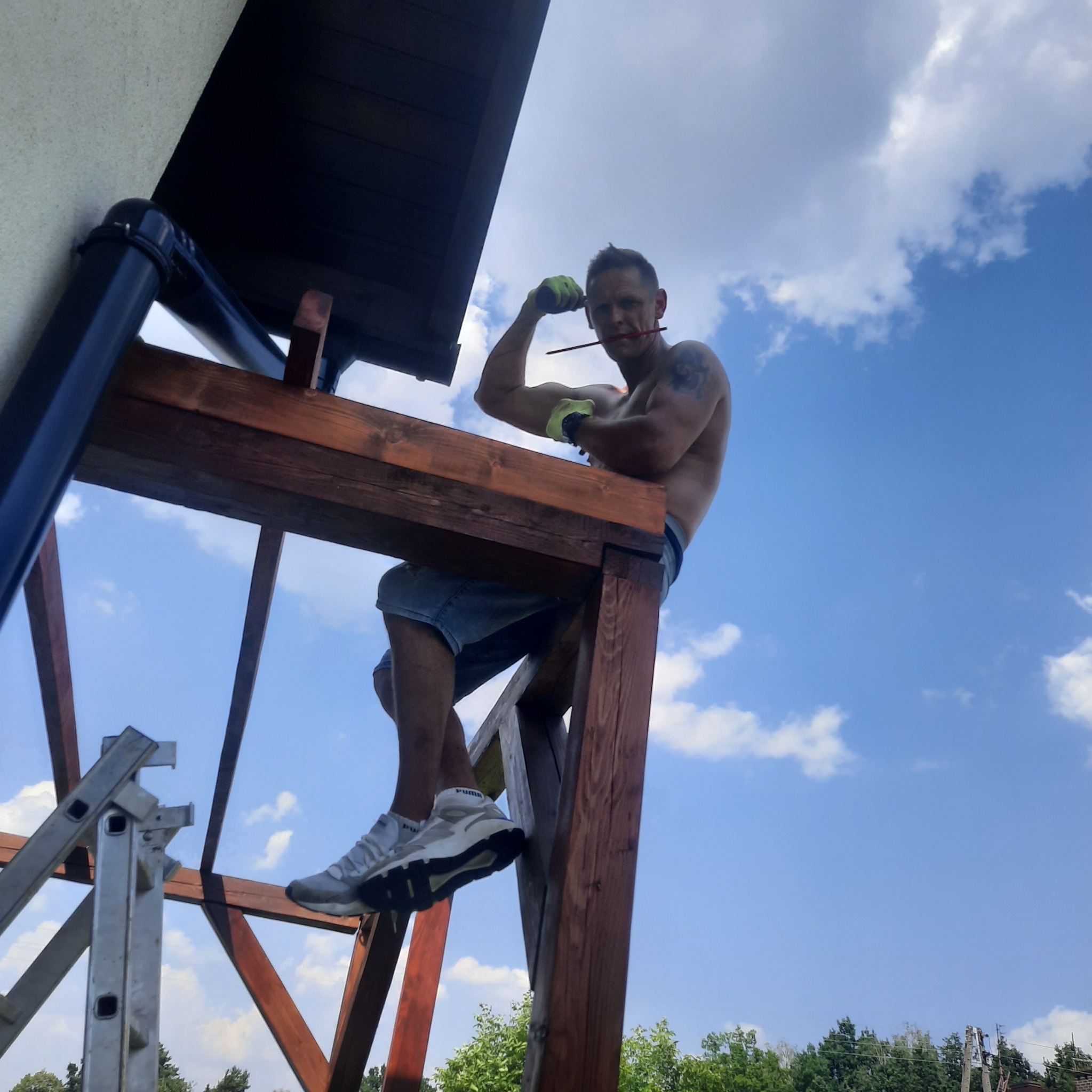 Mężczyzna bez koszulki z tatuażem na ramieniu, w rękawicach roboczych i z ołówkiem w ustach, siedzi na drewnianej konstrukcji pod dachem, w tle błękitne niebo z chmurami.