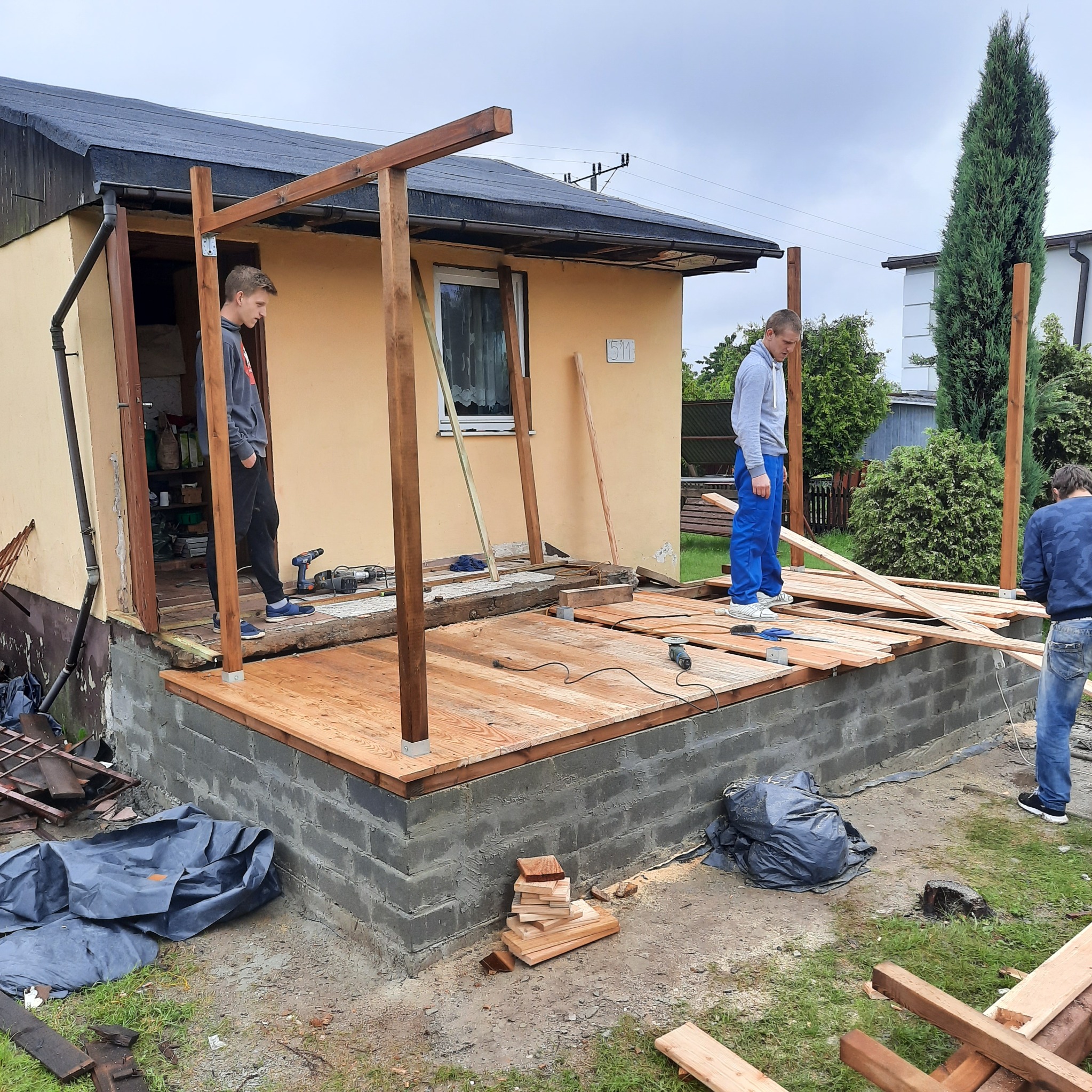 Budowa zadaszonego tarasu: ekipa montuje drewnianą podłogę i konstrukcję nośną z belek, widoczne narzędzia i materiały budowlane, dom z numerem 511 w tle.