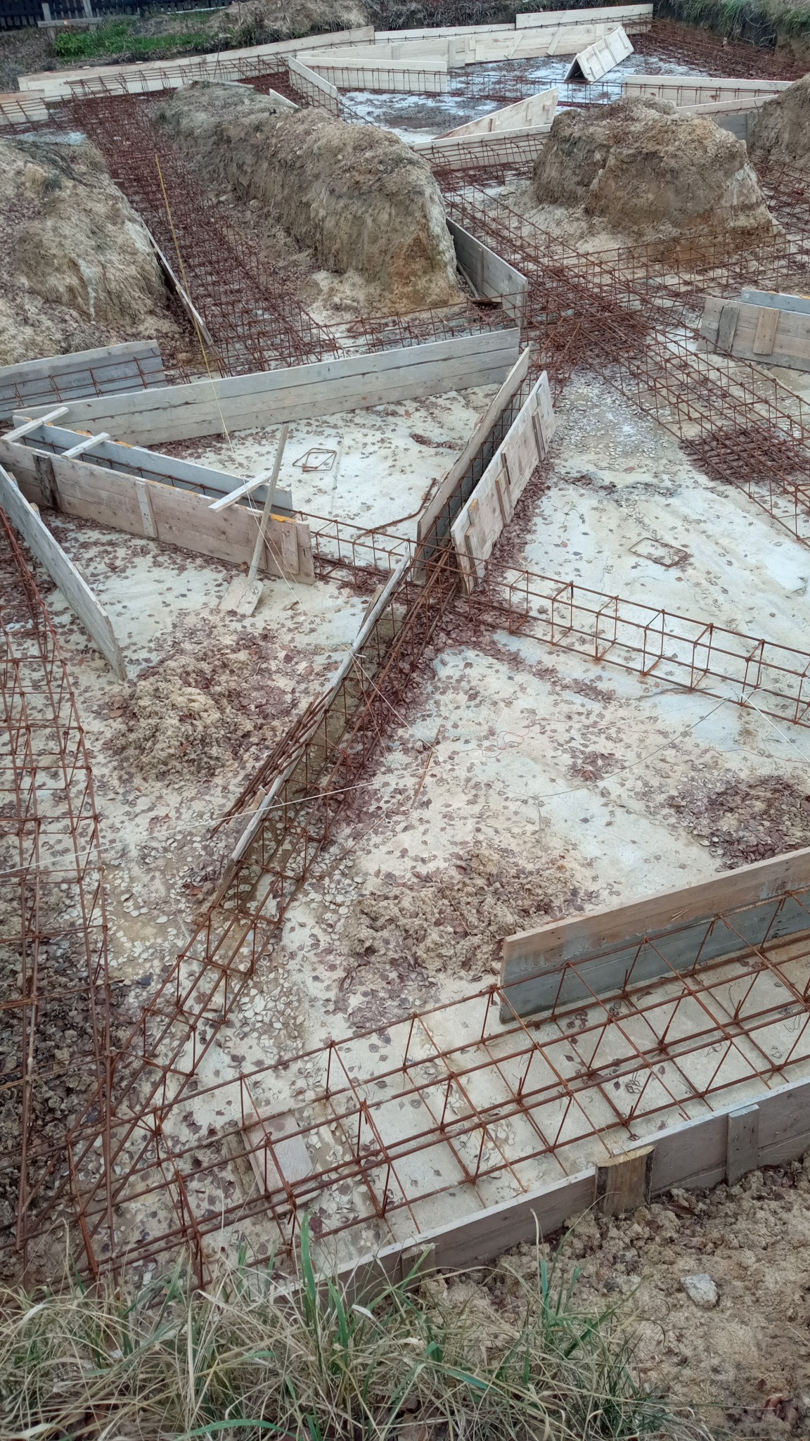 Przygotowanie fundamentów budynku: drewniane szalunki i zbrojenie stalowe na wykopie, widok z góry.