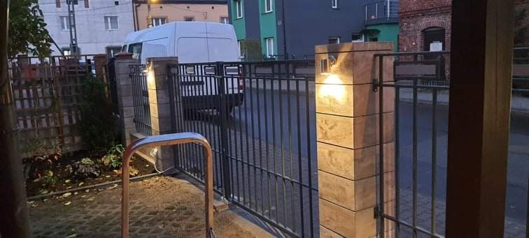 Nowoczesne, metalowe ogrodzenie z kamiennymi słupkami oświetlonymi lampami, widoczne z perspektywy ulicy, z samochodem dostawczym w tle.