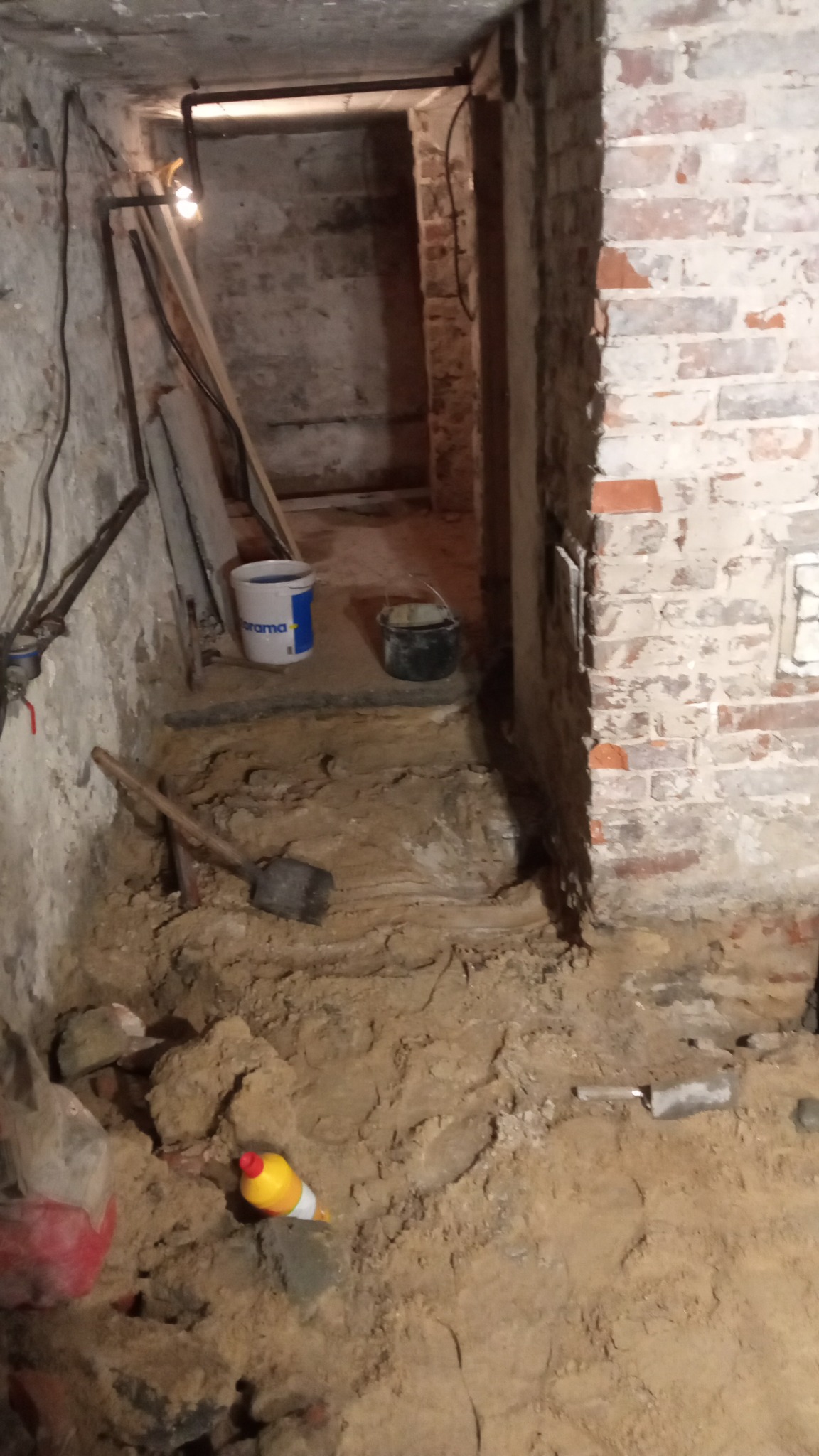 Wykopana ziemia w piwnicy, widoczna łopata, wiadro i butelka z płynem, ściany z cegły i betonu, słabe oświetlenie.