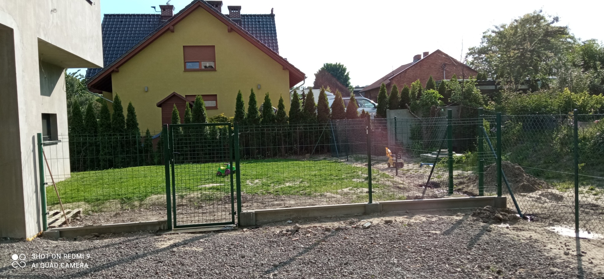 Zielone, metalowe ogrodzenie panelowe z furtką, częściowo zamontowane, widoczne słupki i podmurówka, w tle domy i tuje, na trawniku zabawka dla dziecka.