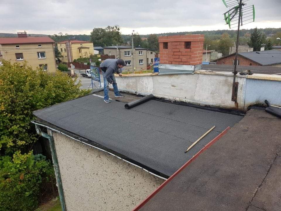 Pracownik w szarej kurtce i jeansach rozkłada czarny pas papy na płaskim dachu budynku z ceglanym kominem i anteną telewizyjną w tle zabudowy miejskiej.