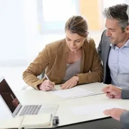 Kobieta podpisuje dokument przy biurku z laptopem, konsultanci obserwują, jasne wnętrze biura.