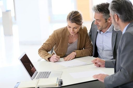 Kobieta podpisuje dokument przy biurku z laptopem, konsultanci obserwują, jasne wnętrze biura.