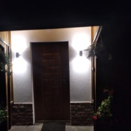 Łukasz USŁUGI BUDOWLANE - Wejście do domu z drewnianymi elementami, oświetlone dwoma lampami ściennymi, z dekoracyjną okładziną z imitacji kamienia u dołu ściany i kwiatami w donicach po bokach.