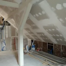 Poddasze w trakcie wykańczania płytami karton gips z widocznymi łatami na łączeniach, drewnianymi belkami konstrukcyjnymi, instalacją elektryczną oraz materiałami budowlanymi w tle.