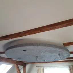 Wykończony sufit z okrągłą, betonową dekoracją w loftowym stylu, z widocznymi drewnianymi belkami stropowymi i otworami na oświetlenie.