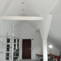 Białe, świeżo pomalowane poddasze z widocznym rusztowaniem, brązowymi drzwiami i lampą budowlaną na statywie, przygotowane do dalszych prac wykończeniowych.