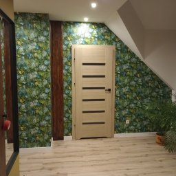 Łukasz USŁUGI BUDOWLANE - Wnętrze po remoncie: Jasne drewniane drzwi z poziomymi wstawkami na tle ściany w tropikalne liście, drewniane panele dekoracyjne, podłoga z paneli imitujących drewno, doniczkowe rośliny w tle.