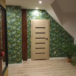 Wnętrze po remoncie: Jasne drewniane drzwi z poziomymi wstawkami na tle ściany w tropikalne liście, drewniane panele dekoracyjne, podłoga z paneli imitujących drewno, doniczkowe rośliny w tle.