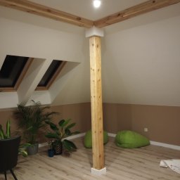 Łukasz USŁUGI BUDOWLANE - Poddasze po remoncie z drewnianą kolumną i belkami stropowymi, oknami dachowymi, roślinami doniczkowymi i zielonymi pufami.