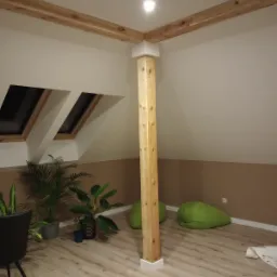 Poddasze po remoncie z drewnianą kolumną i belkami stropowymi, oknami dachowymi, roślinami doniczkowymi i zielonymi pufami.