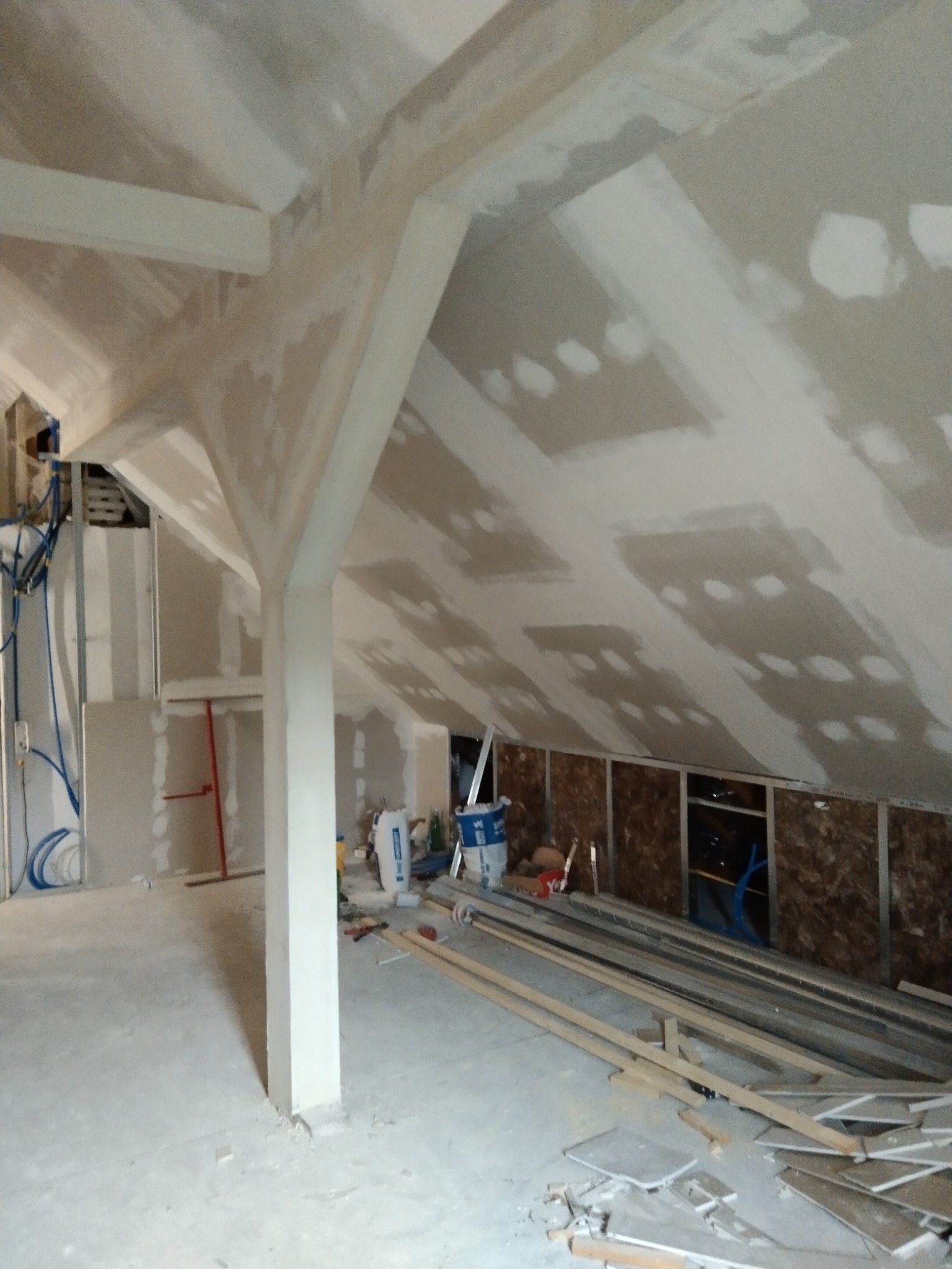 Poddasze w trakcie wykańczania płytami karton gips z widocznymi łatami na łączeniach, drewnianymi belkami konstrukcyjnymi, instalacją elektryczną oraz materiałami budowlanymi w tle.