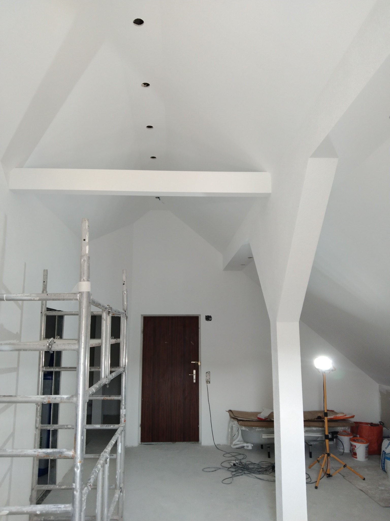 Białe, świeżo pomalowane poddasze z widocznym rusztowaniem, brązowymi drzwiami i lampą budowlaną na statywie, przygotowane do dalszych prac wykończeniowych.