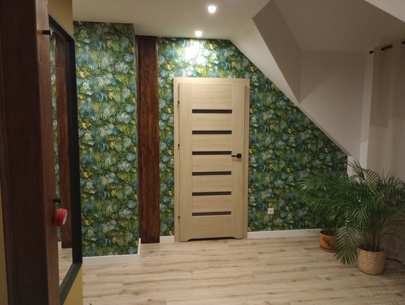 Wnętrze po remoncie: Jasne drewniane drzwi z poziomymi wstawkami na tle ściany w tropikalne liście, drewniane panele dekoracyjne, podłoga z paneli imitujących drewno, doniczkowe rośliny w tle.