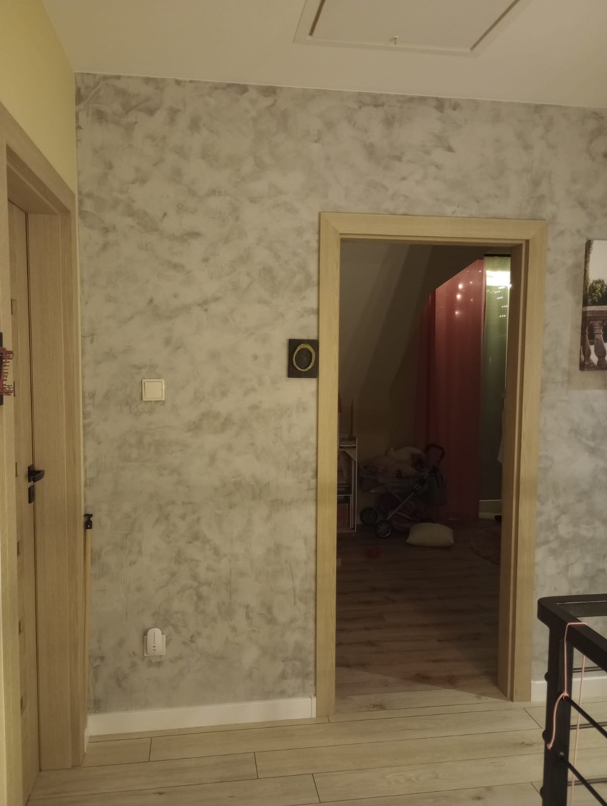 Teksturowana ściana w odcieniach szarości z drewnianymi drzwiami po lewej i ościeżnicą po prawej, widoczny włącznik światła i gniazdko elektryczne, w tle pokój z wózkiem dziecięcym.