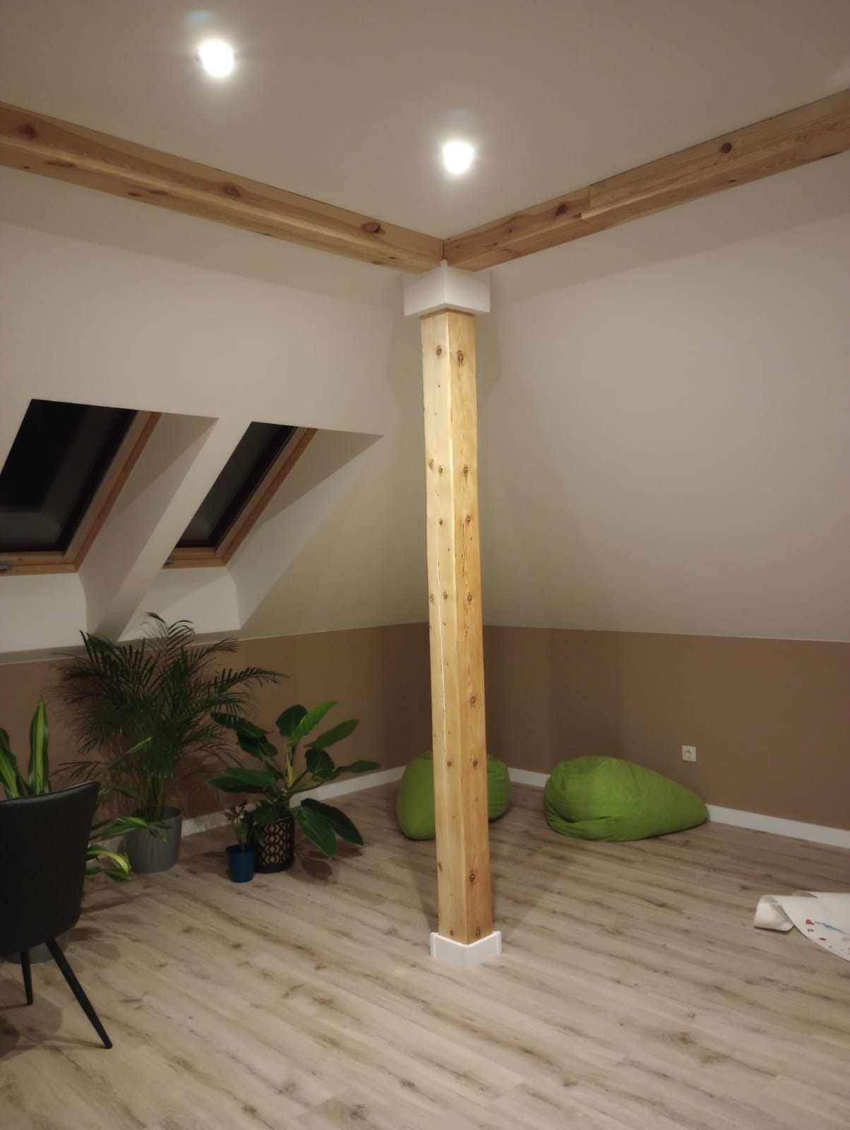 Poddasze po remoncie z drewnianą kolumną i belkami stropowymi, oknami dachowymi, roślinami doniczkowymi i zielonymi pufami.