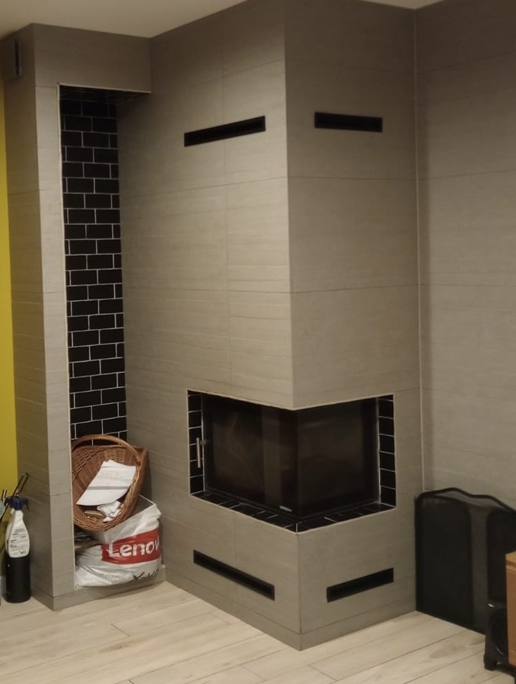 Nowoczesny kominek narożny obłożony szarymi płytkami z czarnymi akcentami, wnęka obok z czarną cegłą i koszem wiklinowym, widoczny fragment żółtej ściany