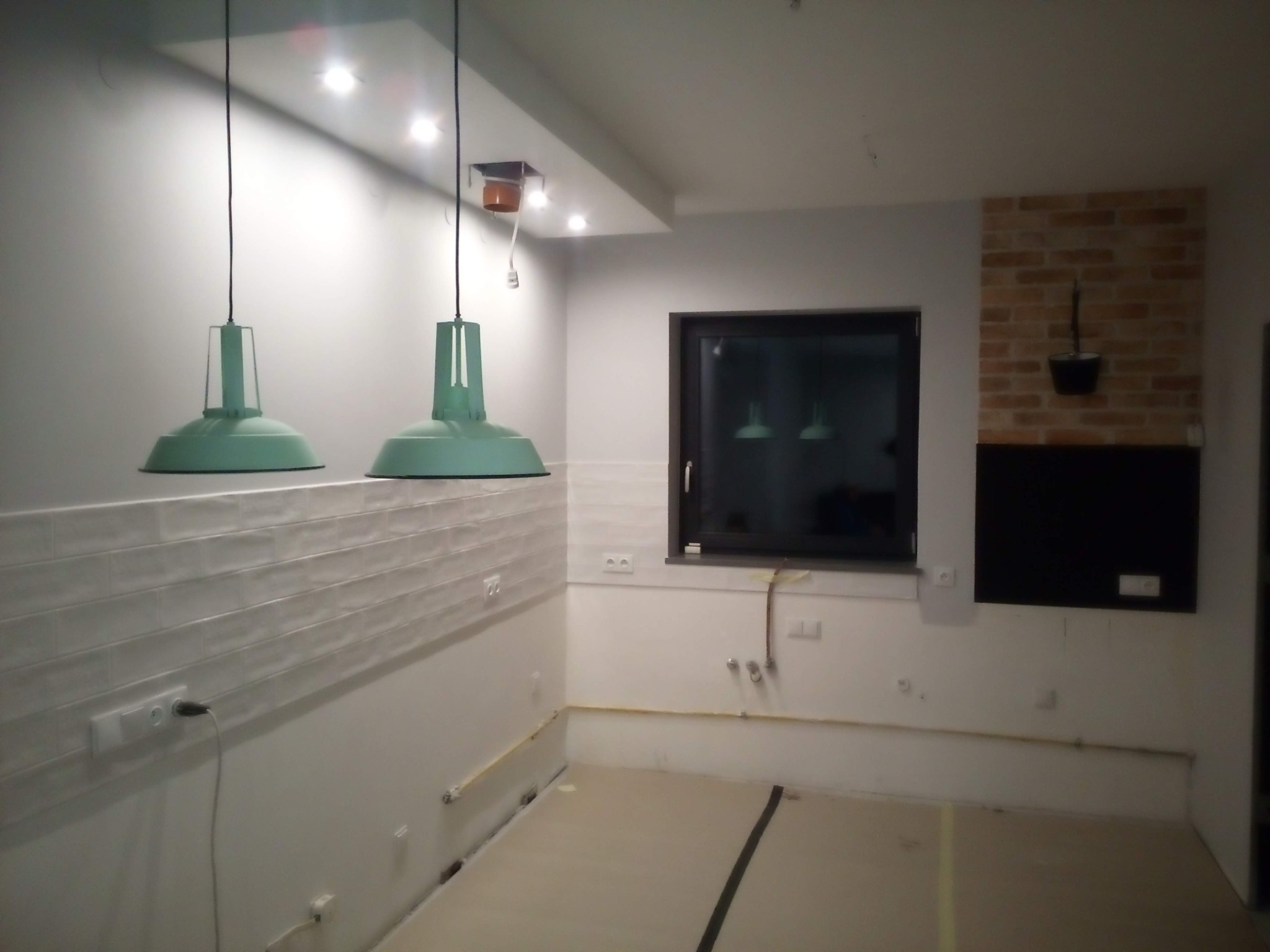 Wnętrze w trakcie remontu: surowe ściany z częściowo położonymi białymi płytkami, widoczne instalacje elektryczne i hydrauliczne, podłoga przygotowana do wykończenia, wiszące lampy w stylu...