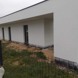 Mg-Bud - Nowoczesny, parterowy dom z płaskim dachem, świeżo po wykończeniu elewacji w kolorze białym. Widoczne czarne ramy okienne i siatka przy gruncie. Ujęcie zza ogrodzenia.