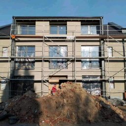 Mg-Bud - Trzy kondygnacyjny budynek w trakcie prac elewacyjnych, osłonięte okna, rusztowania, sterta gruzu na pierwszym planie, słoneczny dzień.