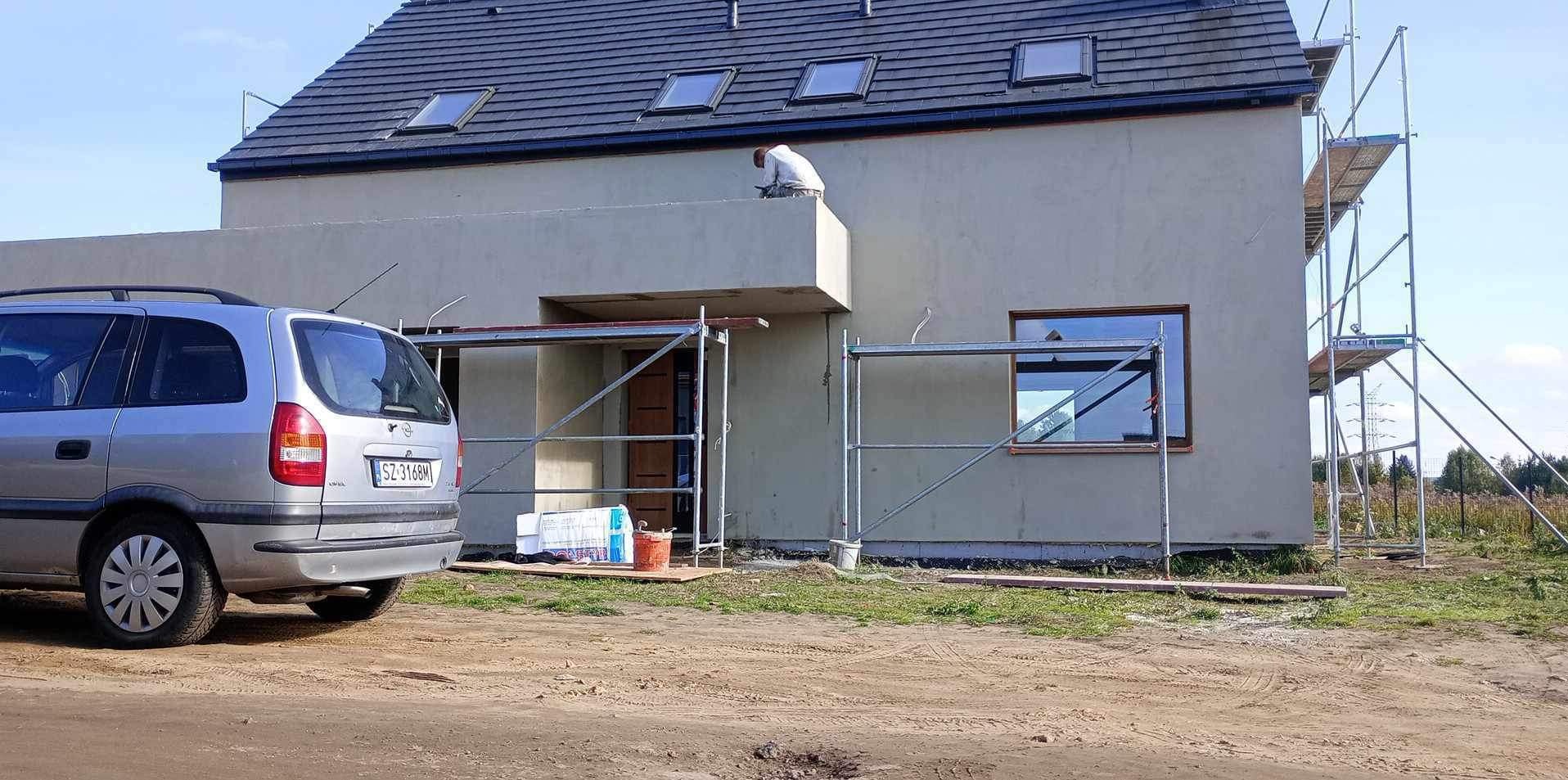 Prace elewacyjne na dwukondygnacyjnym domu z szarą elewacją. Widoczne rusztowania, pracownik na balkonie i zaparkowany samochód osobowy. Ujęcie z dołu.