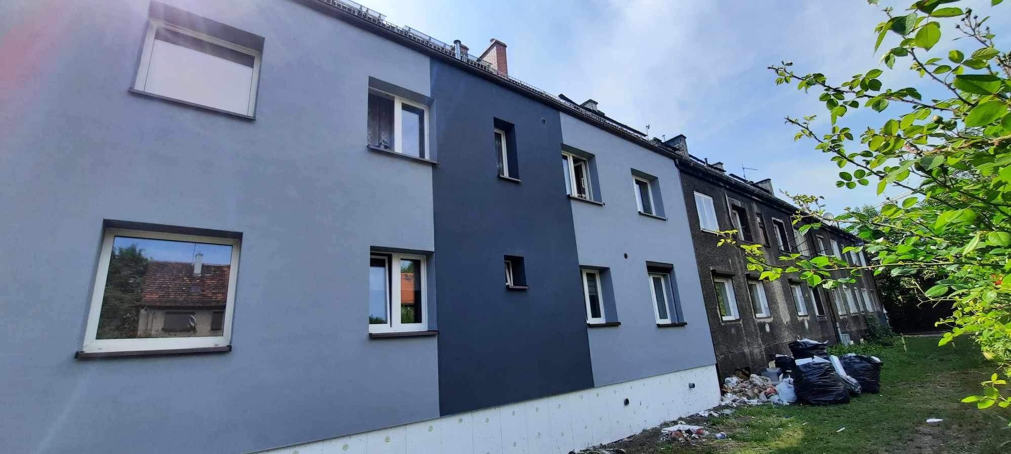 Odnowiona elewacja budynku wielorodzinnego z fragmentem ocieplenia fundamentów, pomalowana na dwa odcienie szarości, sąsiadująca ze starszą, nieodnowioną częścią.
