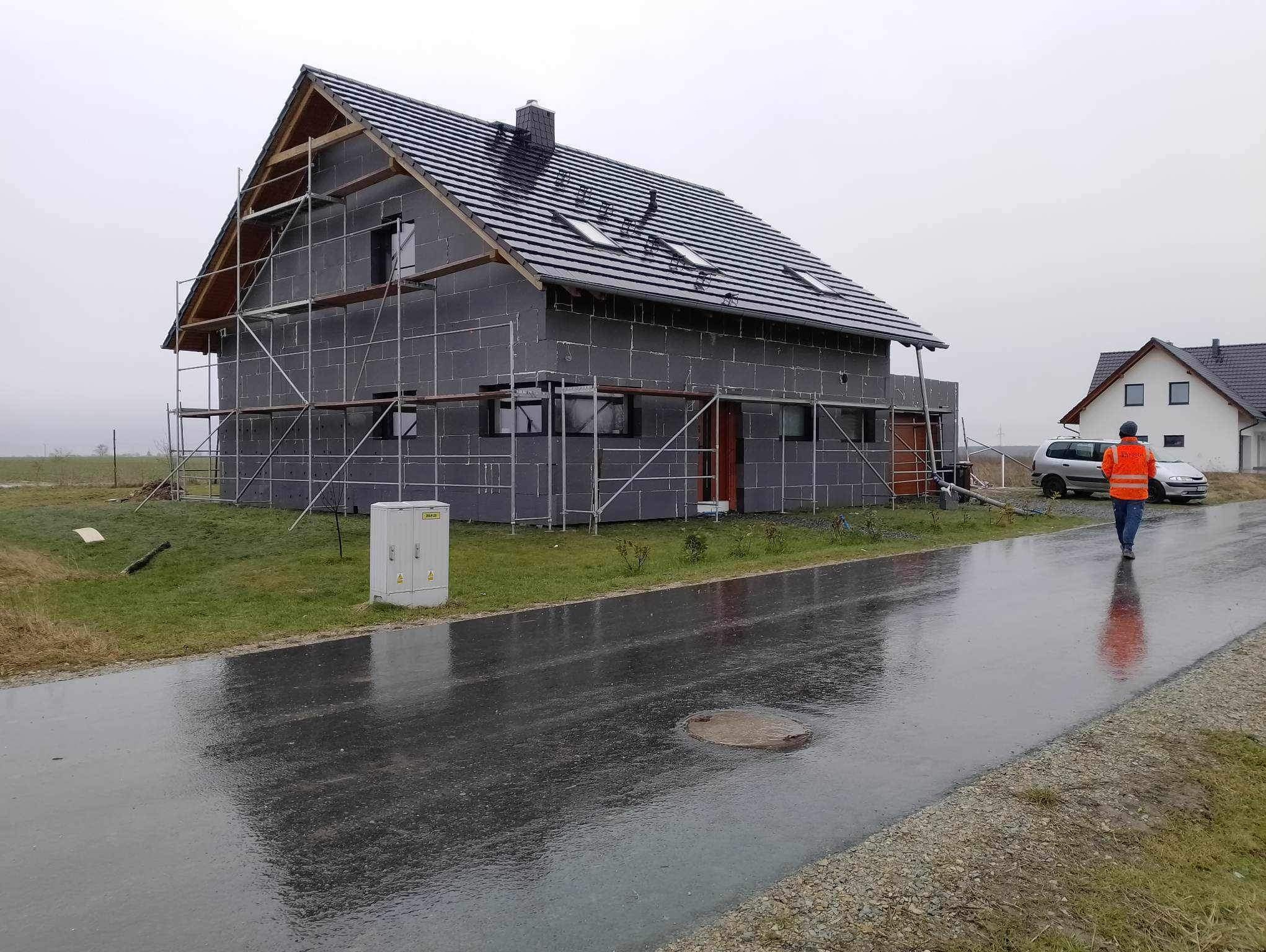 Dom w trakcie ocieplania, pokryty szarymi płytami izolacyjnymi, otoczony rusztowaniami. Na pierwszym planie mokra ulica i idąca osoba w pomarańczowej kamizelce.