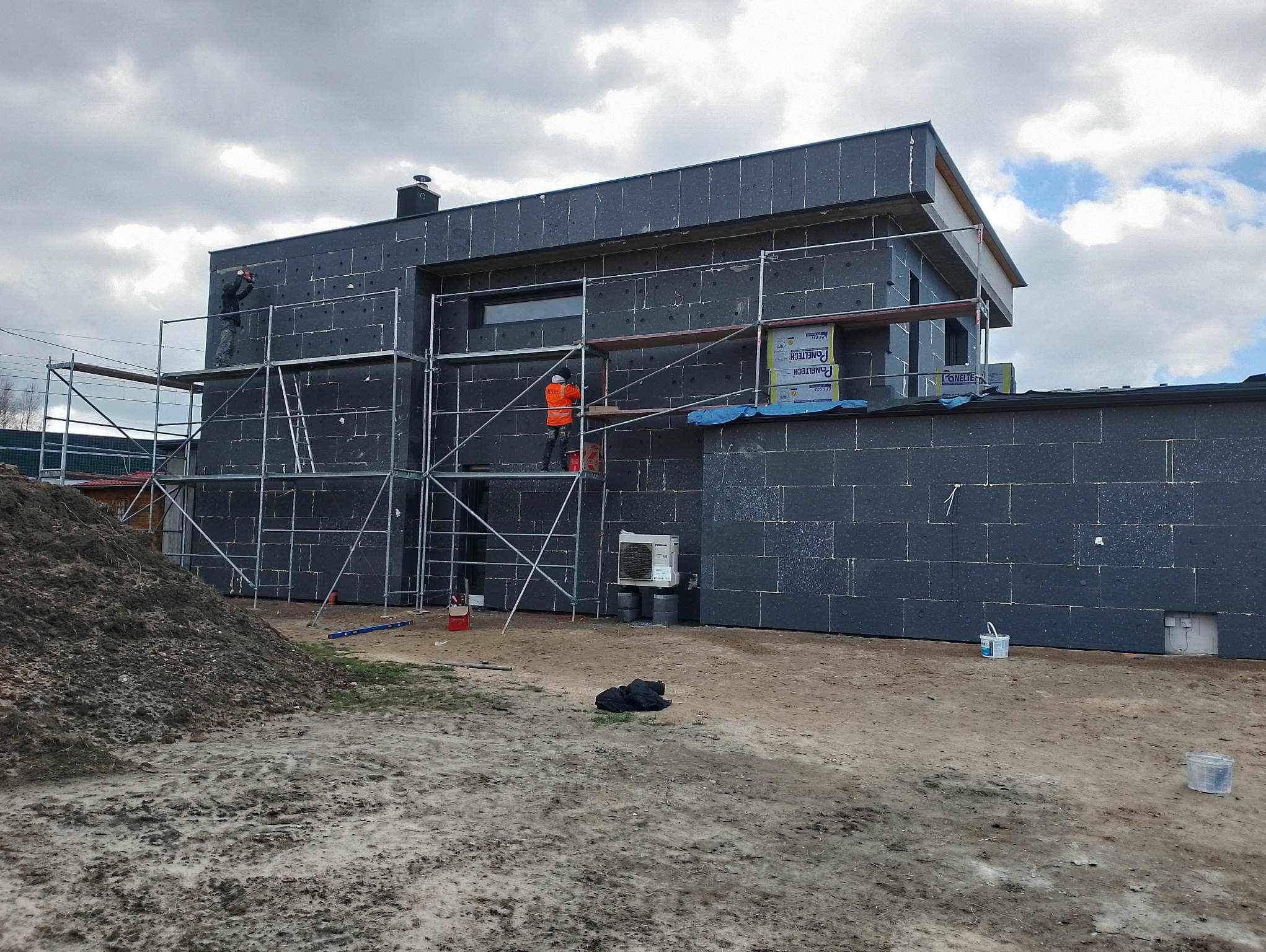 Budowa domu z czarną elewacją, widoczne rusztowania i pracownicy w trakcie montażu izolacji. Na pierwszym planie ziemia i materiały budowlane.