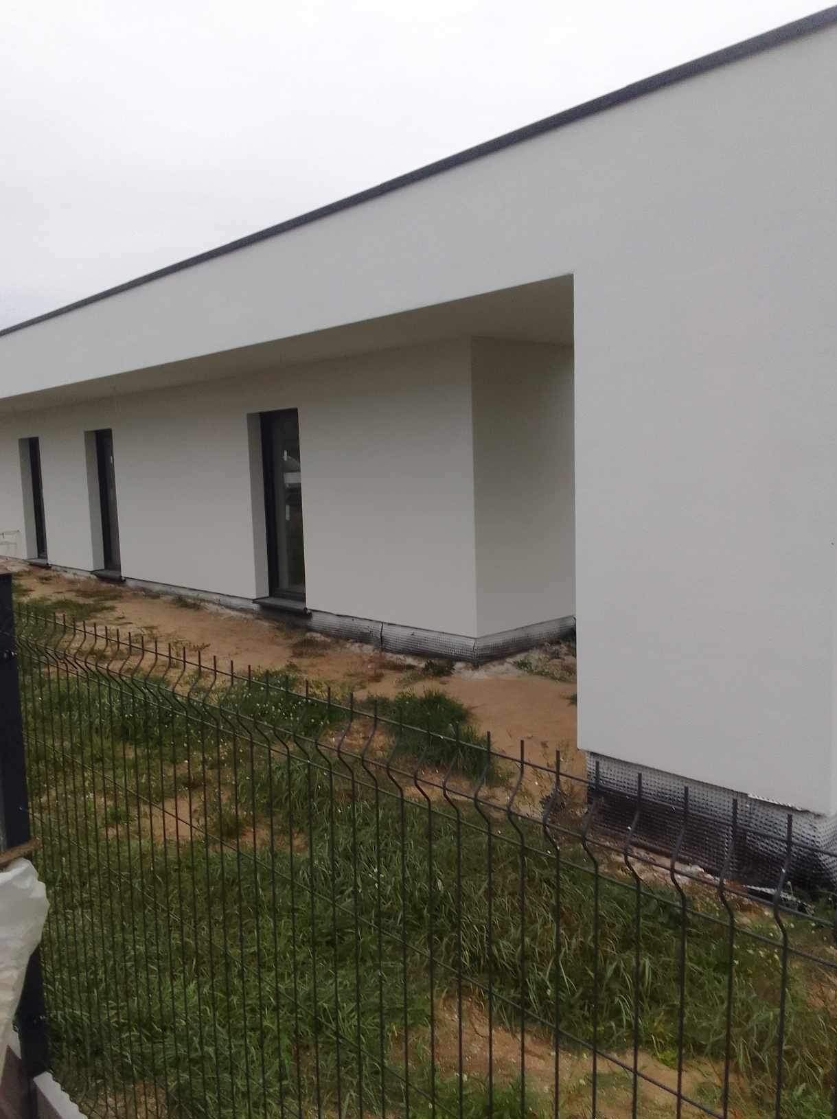 Nowoczesny, parterowy dom z płaskim dachem, świeżo po wykończeniu elewacji w kolorze białym. Widoczne czarne ramy okienne i siatka przy gruncie. Ujęcie zza ogrodzenia.