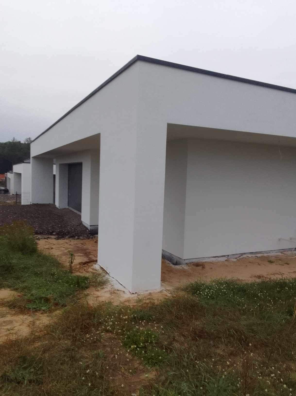 Nowoczesny, parterowy budynek z białymi ścianami i płaskim dachem w trakcie wykończenia. Widoczne elementy elewacji i otoczenie budynku.