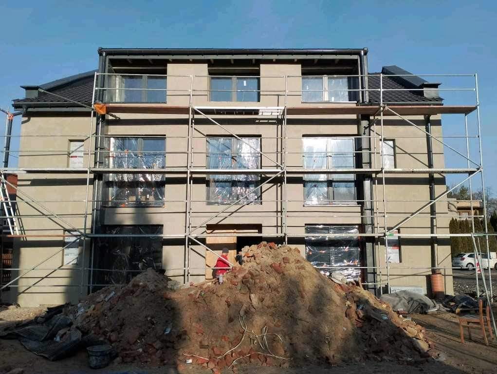 Trzy kondygnacyjny budynek w trakcie prac elewacyjnych, osłonięte okna, rusztowania, sterta gruzu na pierwszym planie, słoneczny dzień.