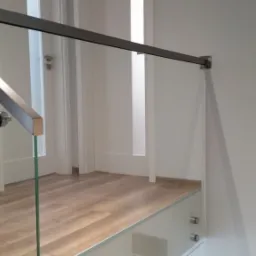 Szklana balustrada z metalowymi elementami mocującymi, widok z boku, minimalistyczny design, jasne wnętrze, drewniana podłoga.