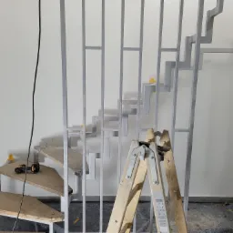 Surowe drewniane schody z metalową konstrukcją w trakcie montażu, widoczna drabina i narzędzia, żółta taśma malarska na konstrukcji.