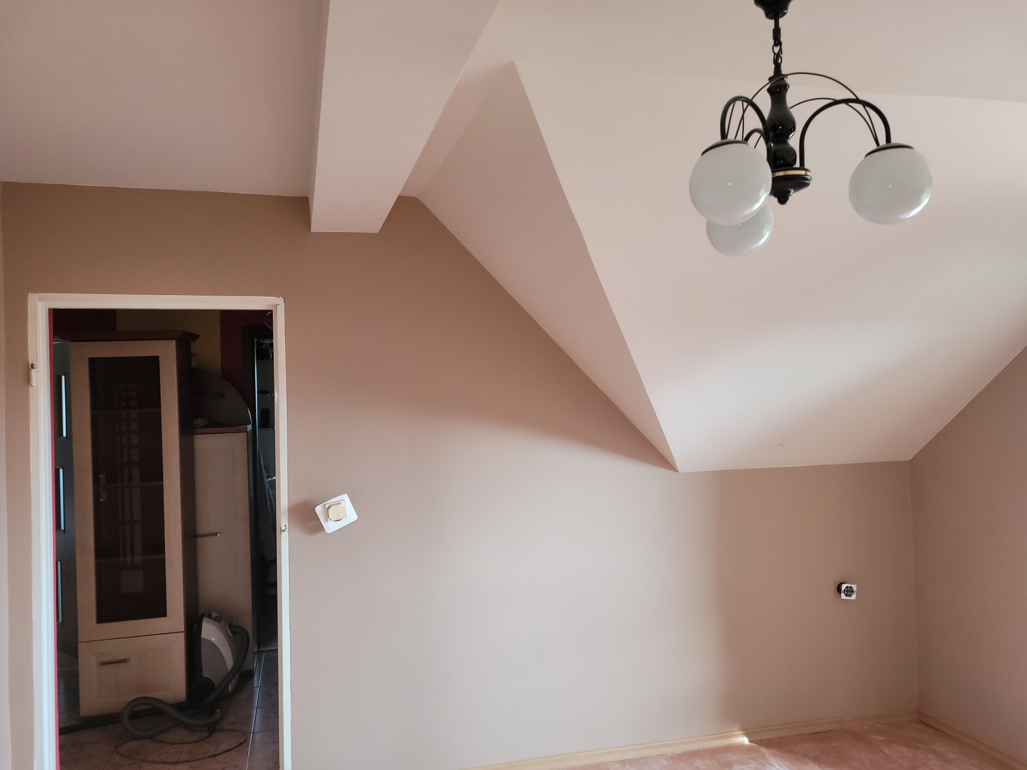 Pomalowane na beżowo poddasze ze skosami, widoczny czarny żyrandol z białymi kulami i gniazdko elektryczne na ścianie, w tle otwarte drzwi do innego pomieszczenia z meblami i odkurzaczem.