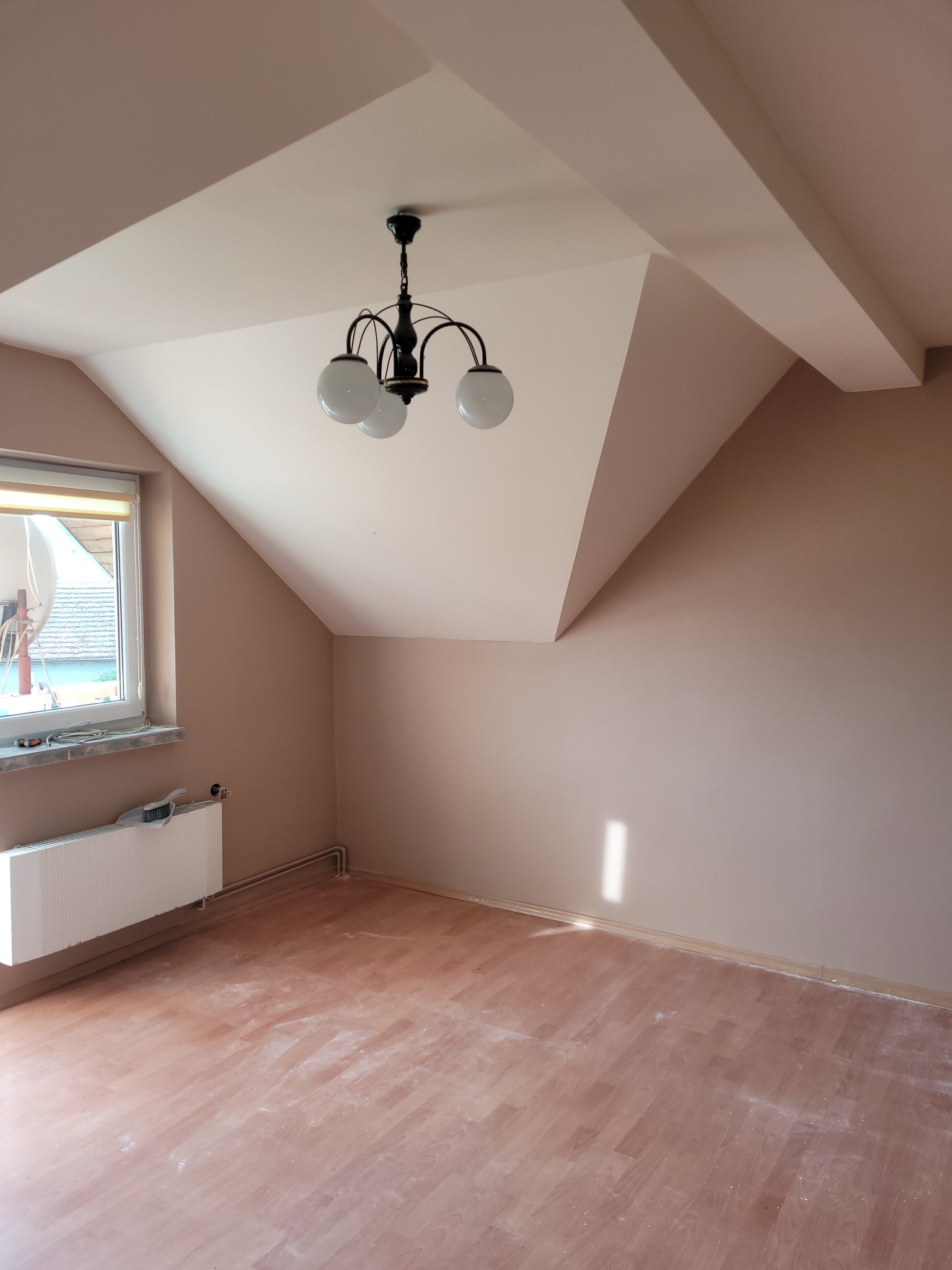 Poddasze po malowaniu ścian na kolor brzoskwiniowy, widoczny biały grzejnik pod oknem, drewnopodobne panele podłogowe z resztkami farby oraz czarny, klasyczny żyrandol.