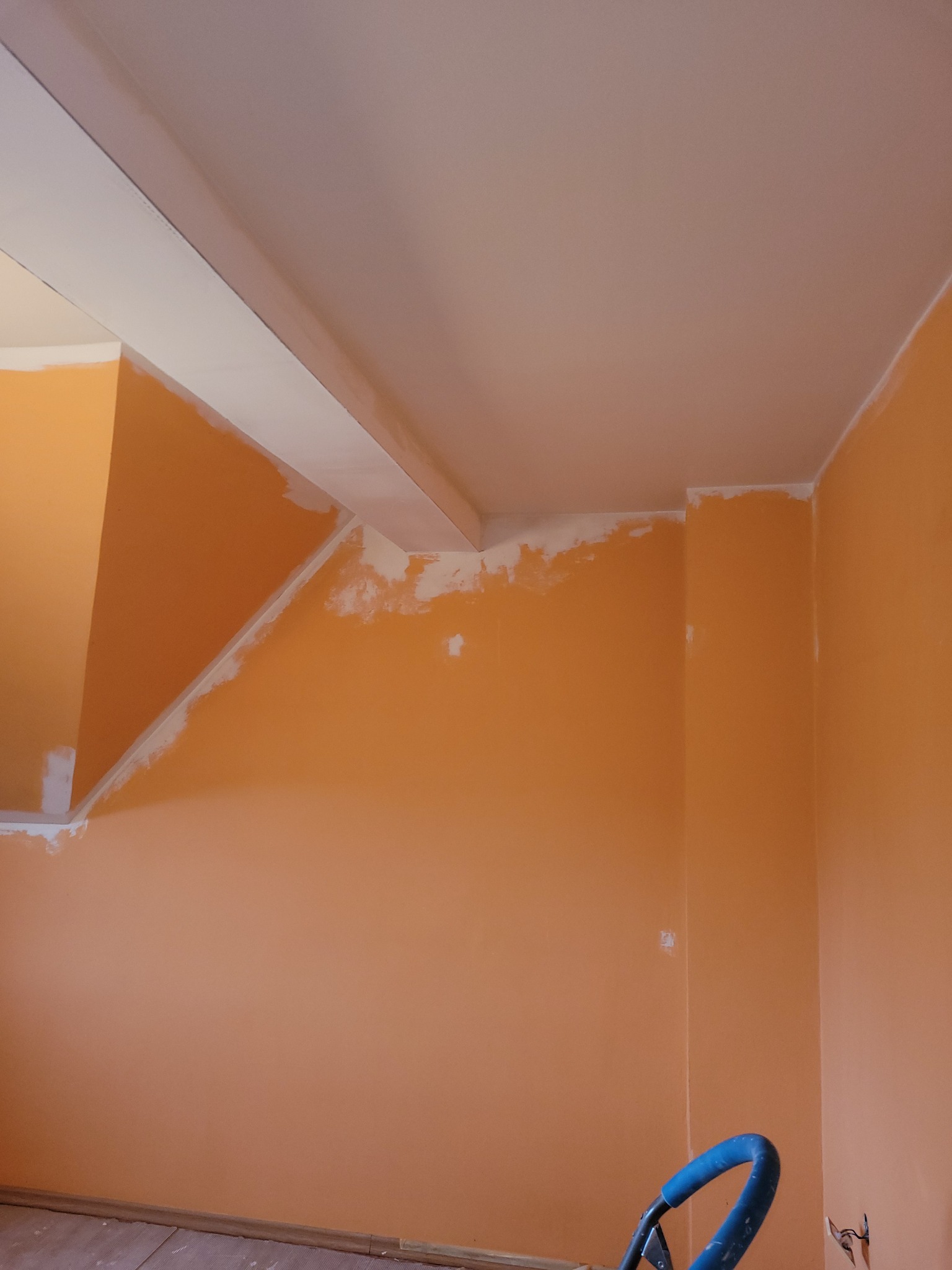 Pokój w trakcie malowania na pomarańczowo, z widocznymi fragmentami świeżo położonej białej szpachli na ścianach i suficie, oraz niebieskim wózkiem malarskim w dolnej części kadru.