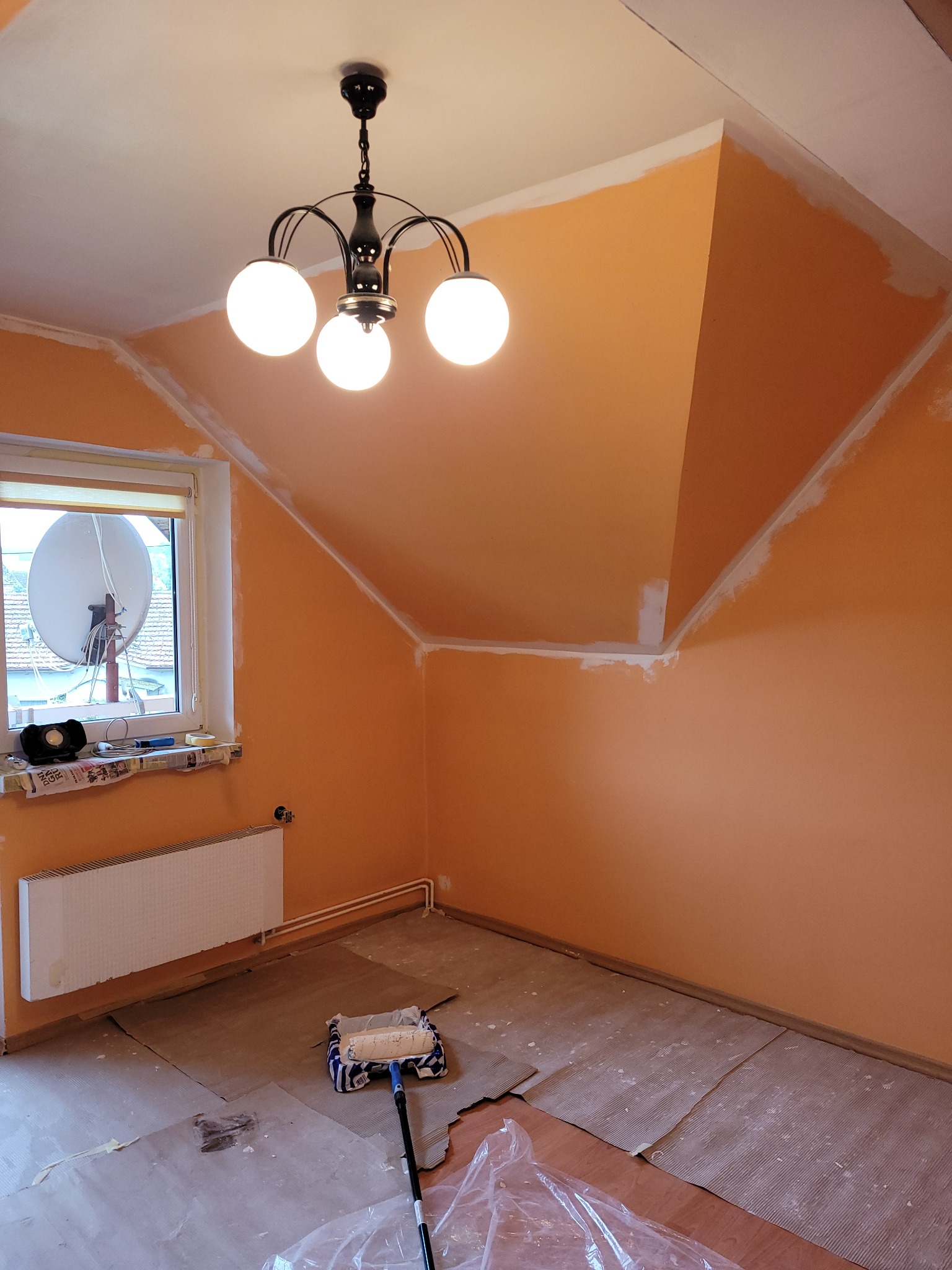 Pomarańczowe wnętrze poddasza w trakcie malowania, z widocznym wałkiem malarskim, osłoniętą podłogą i lampą sufitową z trzema białymi kulami.