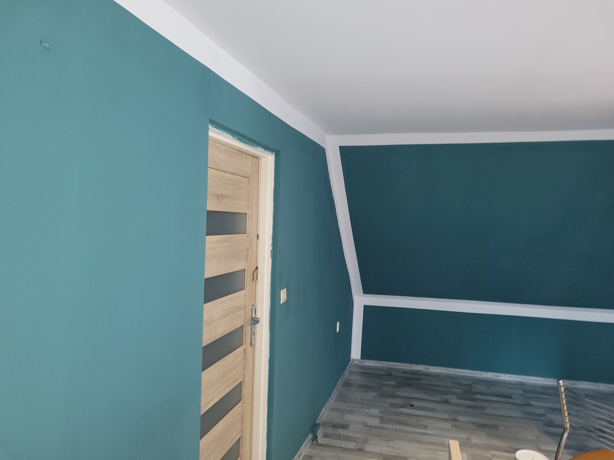 Turkusowe ściany świeżo pomalowanego pokoju z drewnianą stolarką drzwiową, białe listwy wykończeniowe i szara podłoga z paneli, widoczne ślady po malowaniu.
