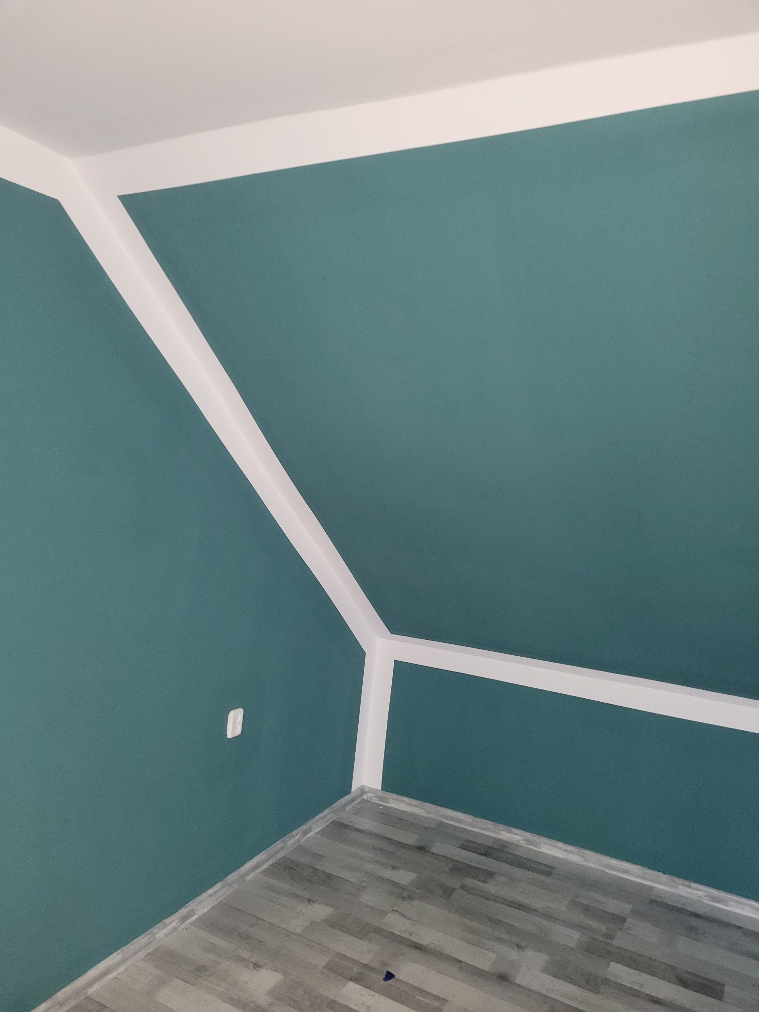 Ściana pomalowana na turkusowo z białym wykończeniem przy suficie w Rudnej, widoczny szary parkiet na podłodze i gniazdko elektryczne.