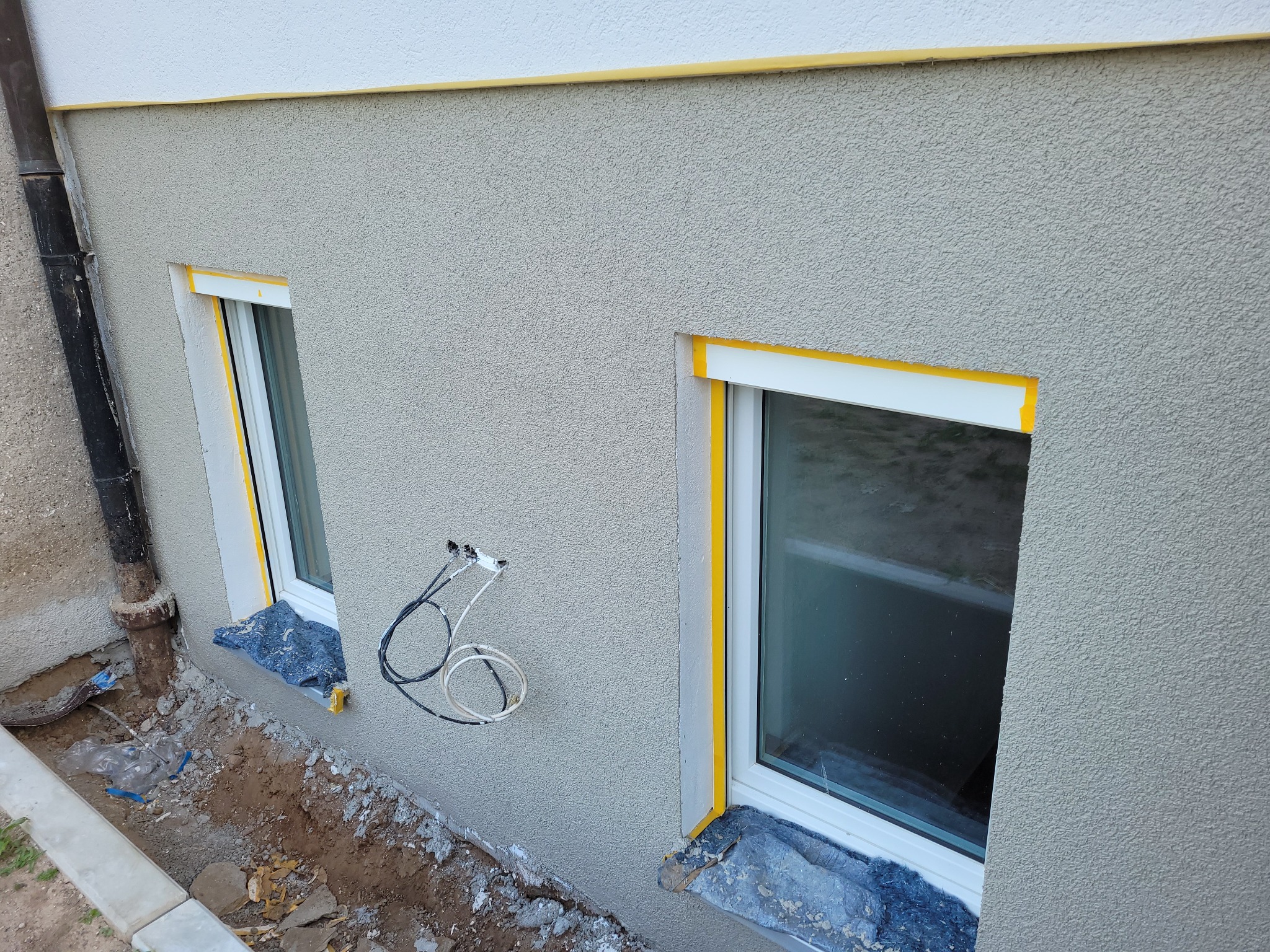 Świeżo otynkowana elewacja budynku z dwoma oknami zabezpieczonymi żółtą taśmą malarską, widoczne przewody elektryczne i rura spustowa, teren wokół okien zabezpieczony folią.