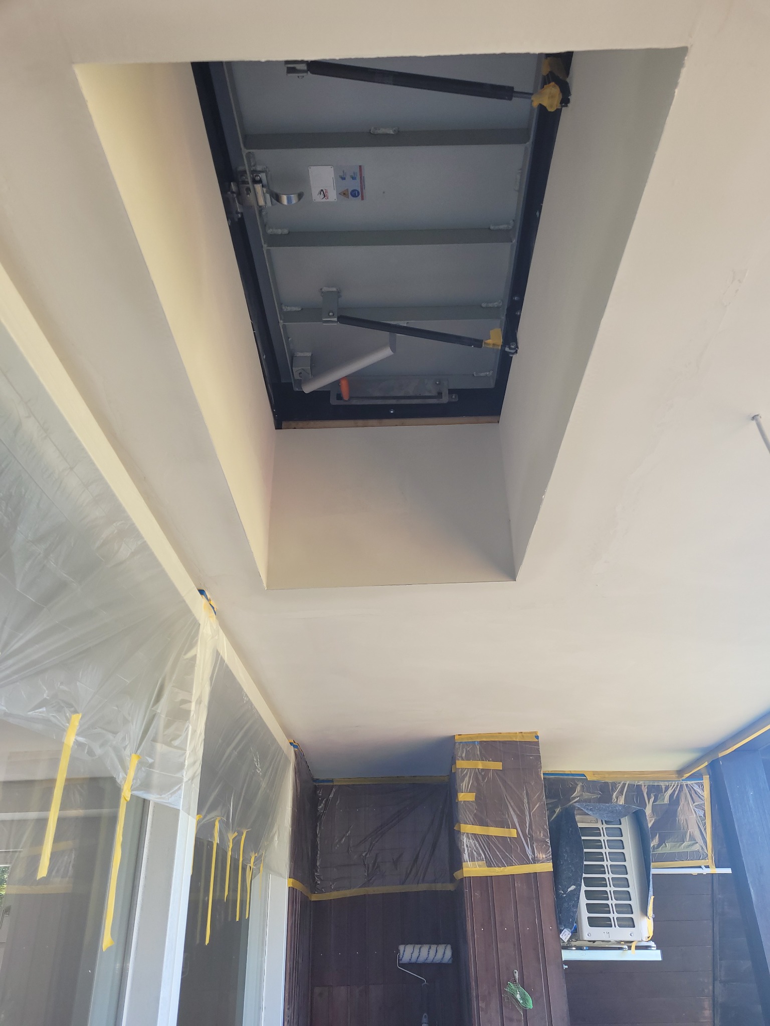 Wnętrze pomieszczenia w trakcie prac malarskich, sufit i ściany pokryte świeżą warstwą farby, zabezpieczone okna i klimatyzator folią malarską, widoczny otwór inspekcyjny w suficie.