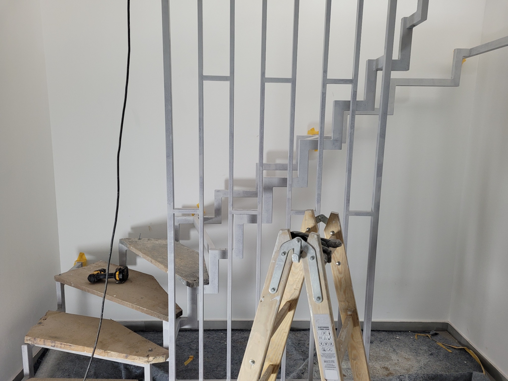 Surowe drewniane schody z metalową konstrukcją w trakcie montażu, widoczna drabina i narzędzia, żółta taśma malarska na konstrukcji.