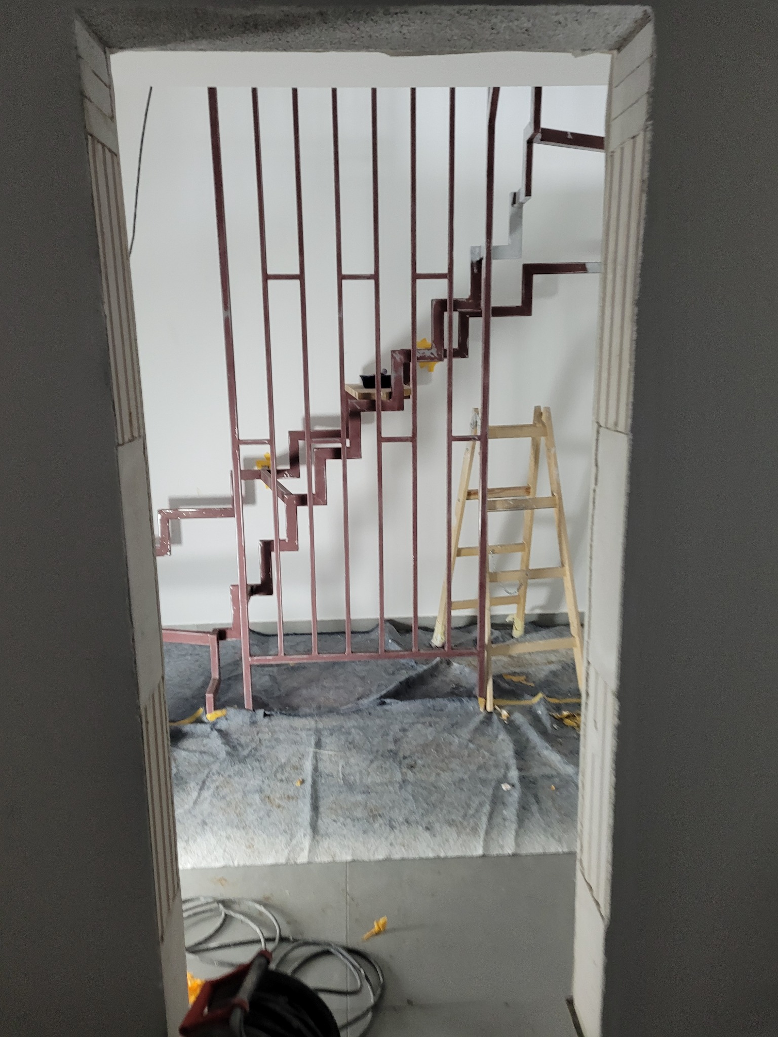 Wnętrze w trakcie remontu z widokiem na metalową konstrukcję schodów, zabezpieczoną podłogę i drabinę, widziane z perspektywy innego pomieszczenia.