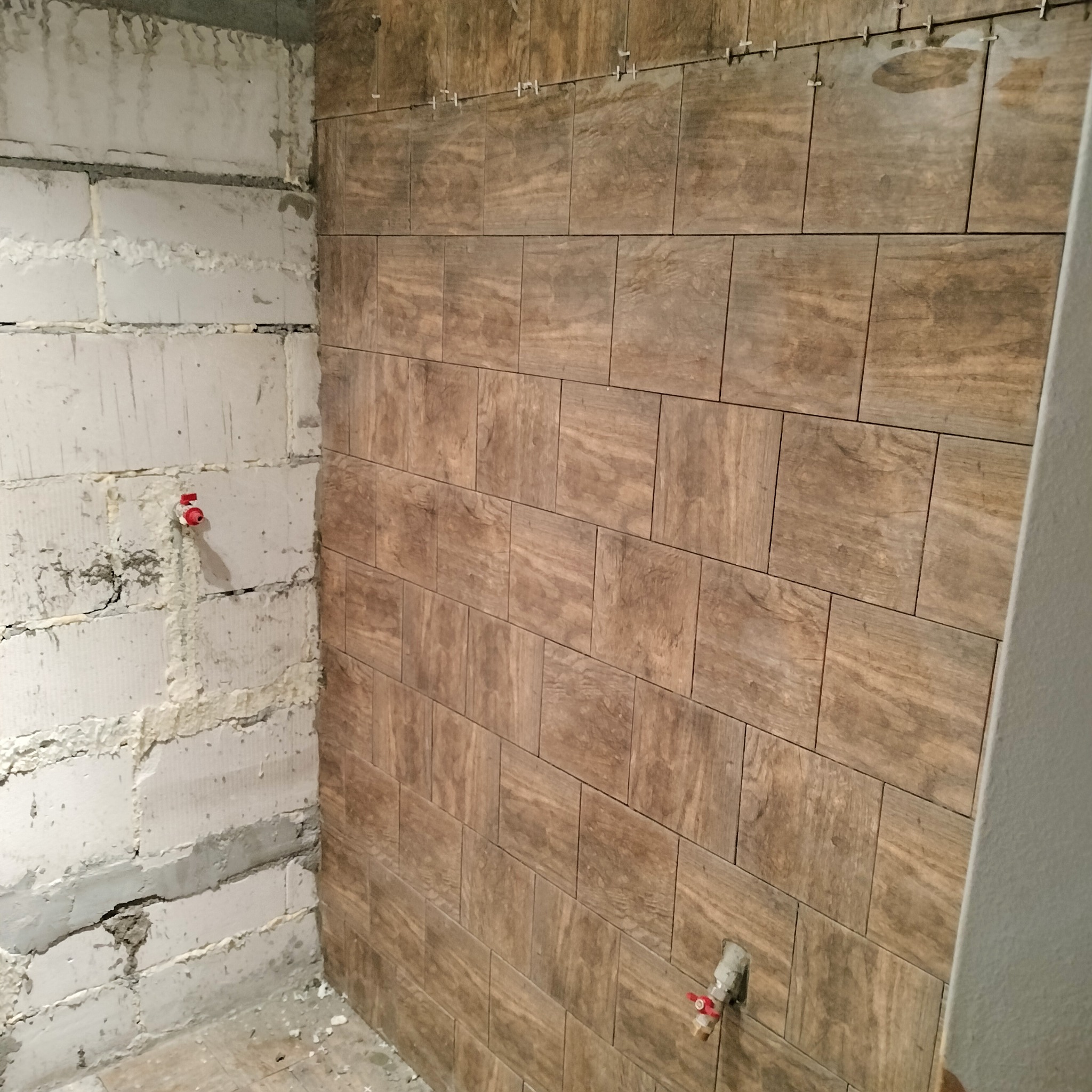 Ściana z brązowych płytek imitujących drewno w trakcie montażu, widoczne krzyżyki dystansowe, surowa ściana z betonu komórkowego z wystającymi rurkami instalacji wodnej.