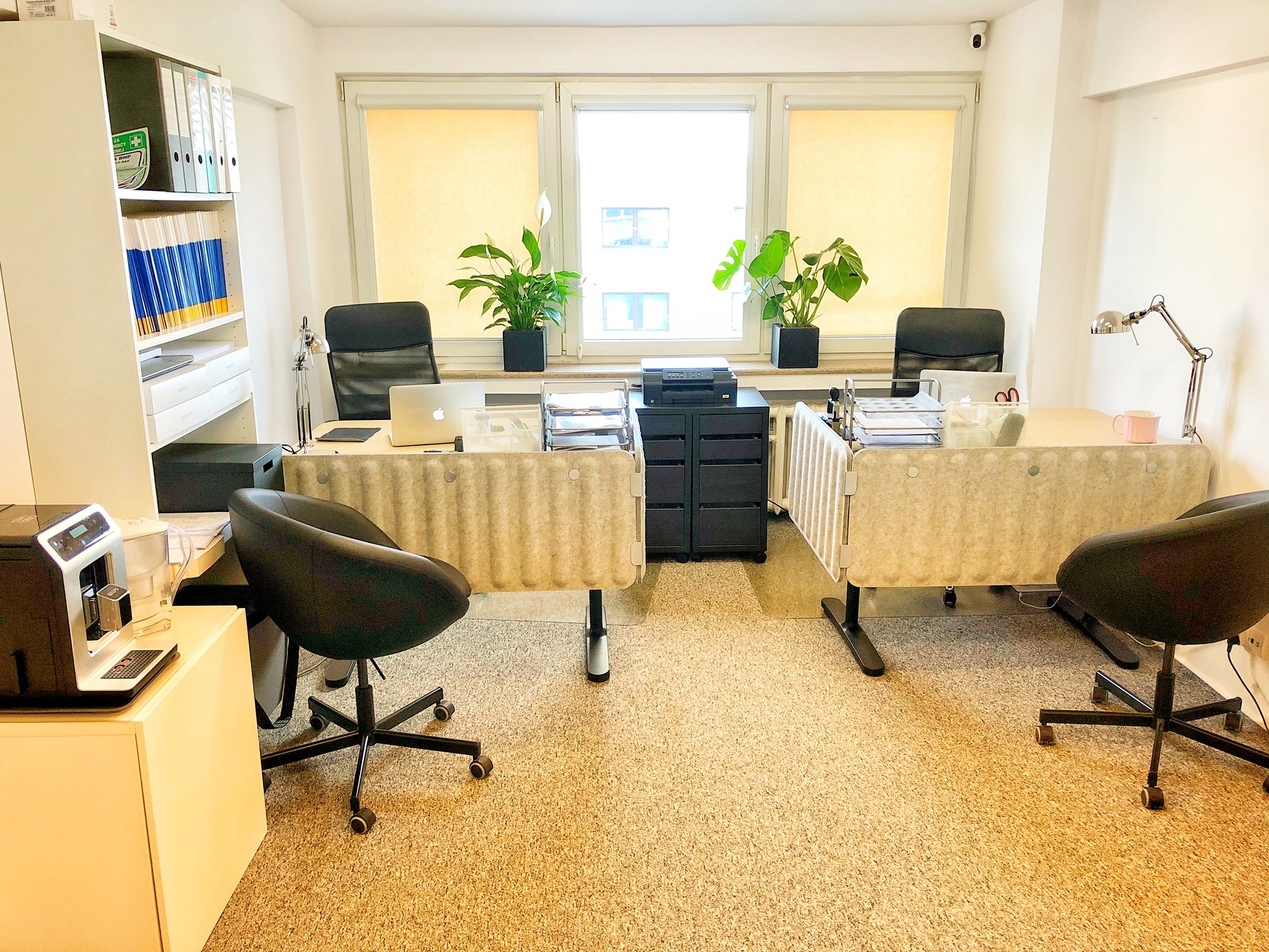 Nowoczesne biuro z dwoma stanowiskami pracy, roślinami doniczkowymi na parapecie, ekspres do kawy i białymi szafkami na dokumenty.